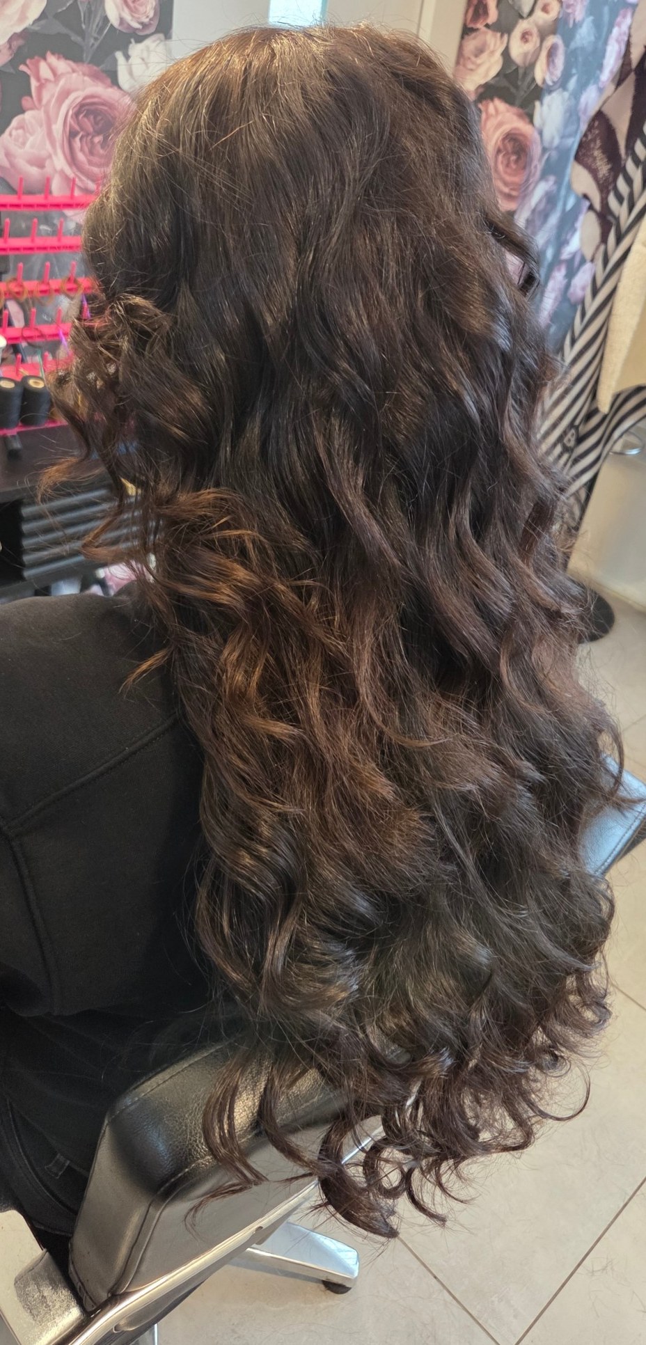 Lange donkere krullen in de salonstoel, met het roze rozenbehang van de salon op de achtergrond